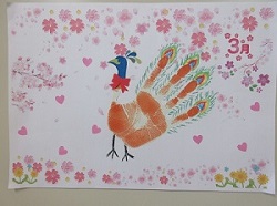 制作する手形カードのイメージ。赤色の手形をクジャクの体や羽根に見立て、顔や足の飾りをつけています。クジャクの周りには桜やハートなどの飾りがあり、隅に「3月」と記載されています。