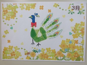 制作する手形カードのイメージ。緑色の手形をクジャクの体や羽根に見立て、顔や足の飾りをつけています。クジャクの周りには黄色の花の飾りがあり、隅に「3月」と記載されています。