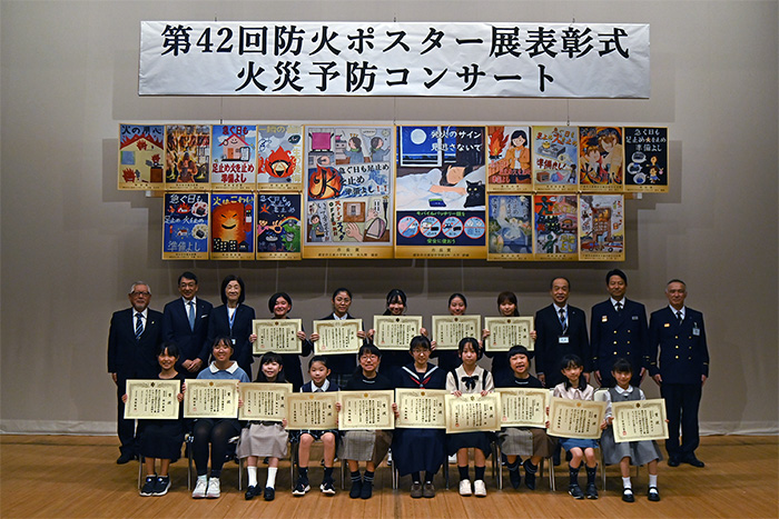 表彰式での記念写真。写真上部には「第42回防火ポスター展表彰式　火災予防コンサート」の看板と、入賞したポスターが掲示されています。写真下部に賞状を持った入賞者と野﨑副市長など関係者が写っています。